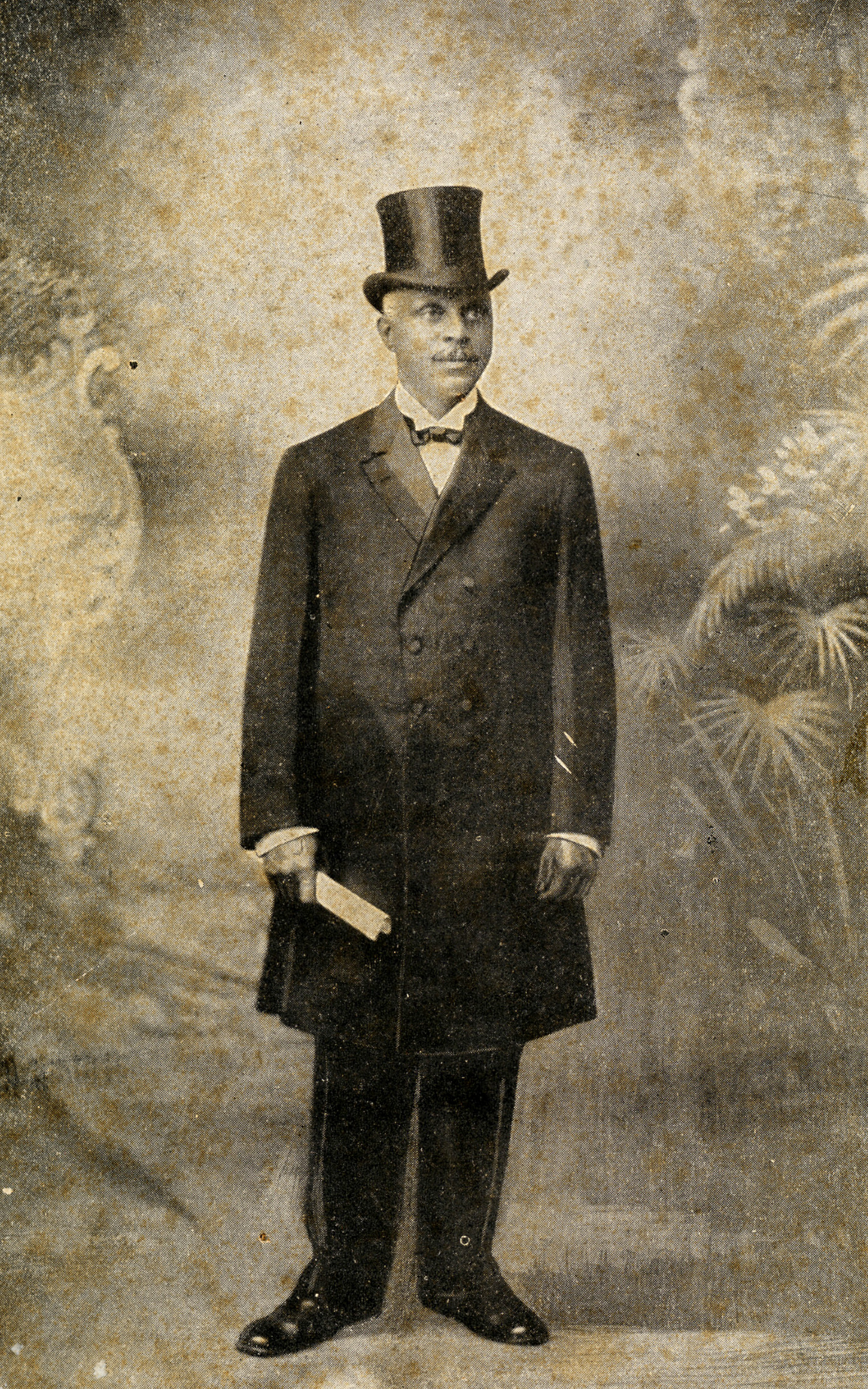 Taylor wearing a dark suit and top hat and holding what looks like a folded paper.