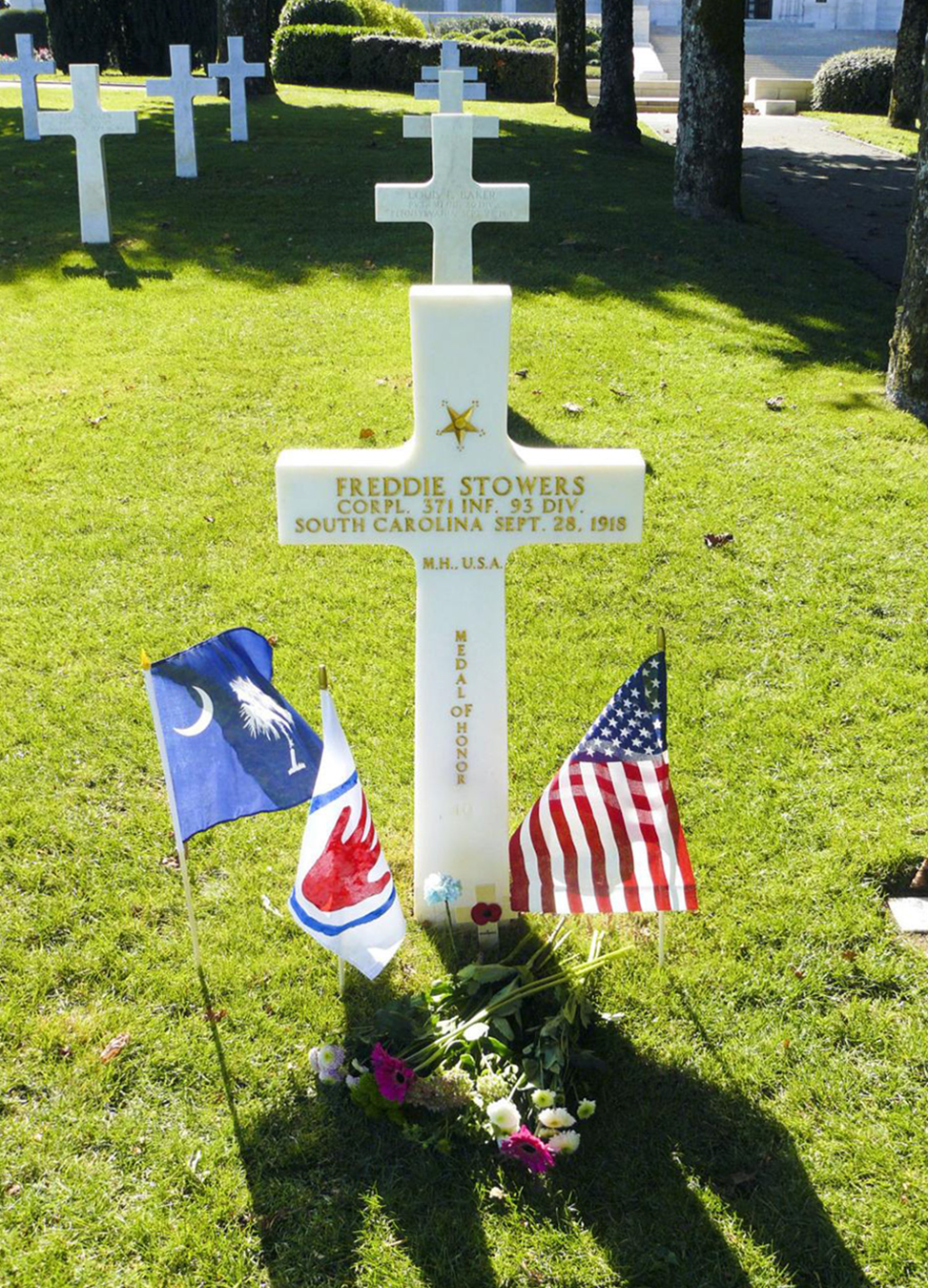 Cross marker with small flags and a bed of flowers in front of it|