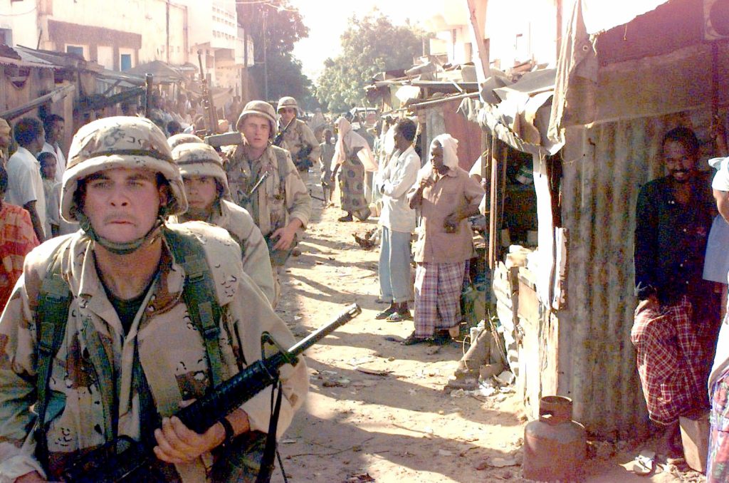 As Somali civilians watch, US Marines walk single file toward the camera, down a small ally in Somalia's Bakara Market. The Marines sweep the market looking for arms and munitions as part of Operation Nutcracker. This mission is in direct support of Operation Restore Hope