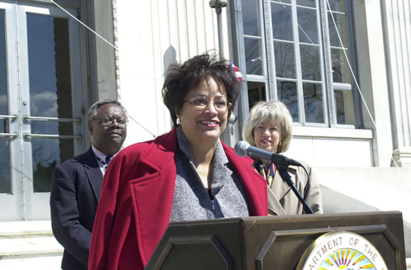 James speaking at podium with two people in the background listening