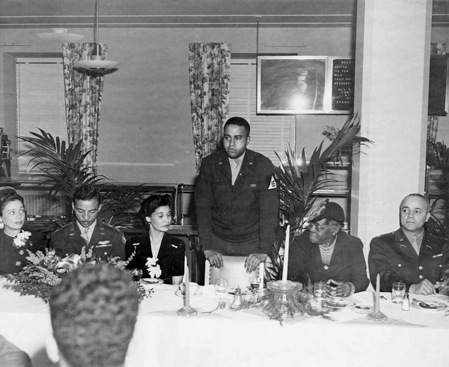 Moss leaning on a chair while speaking as five seated attendees listen|Poster featuring Black soldier wearing a helmet and carrying a rifle with orange background and gold lettering